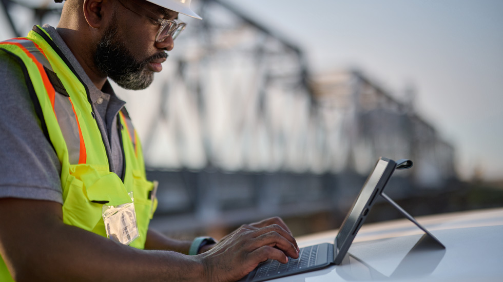 A man wearing a hard hat and typing on an iPad. He is outside and behind him is a bridge.